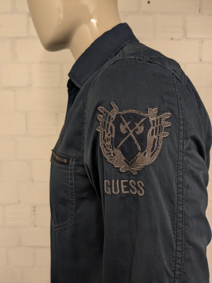 Devinez la chemise en jeans. Bleu foncé coloré avec des accents à fermeture éclair avec des sacs à poitrine. Taille L.