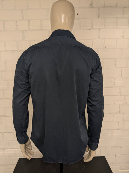 Devinez la chemise en jeans. Bleu foncé coloré avec des accents à fermeture éclair avec des sacs à poitrine. Taille L.