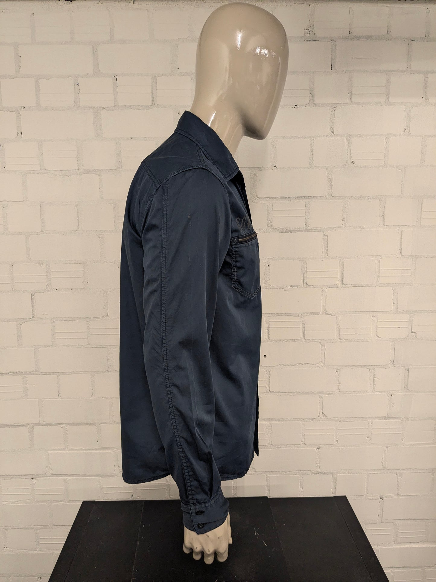 Devinez la chemise en jeans. Bleu foncé coloré avec des accents à fermeture éclair avec des sacs à poitrine. Taille L.